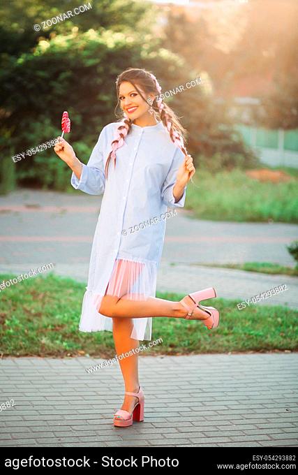 Fashionable positivity girl wearing in blue dress, with pink braids after beauty salon, posing, smiling at camera. Pretty brunet woman holding sweet candy heart...