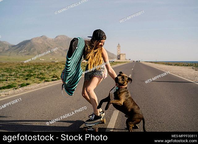 Young woman with surfboard and dog skateboarding on asphalt road, Almeria, Spain