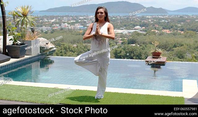 Serene adult concentrated female in white clothes practicing yoga in tree pose poolside against wonderful landscape of green island and sea coast