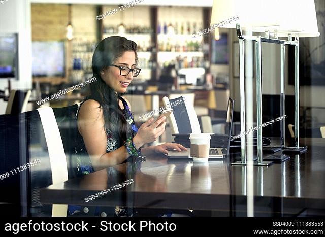 Hispanic woman using cell phone and laptop in coffee shop