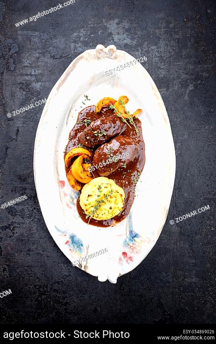 Traditional German braised pork cheeks in brown red wine sauce with fried mushed potatoes and pumpkin as top view on a shabby chic design white plate with copy...
