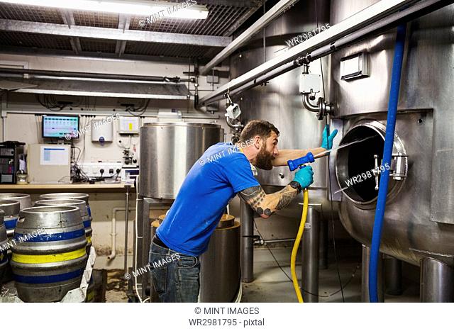 Man working in a brewery, cleaning inside of a large stainless steel kettle with a high pressure washer