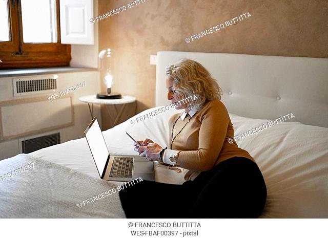 Mature businesswoman lying on bed with laptop using mobile phone