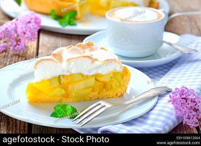 Leckerer Rhabarberkuchen mit Baiserhaube und Cappuccino auf einem Holztisch mit fruehlingshafter mit Fliederdeko - Delicious rhubarb cake with springlike lilac...