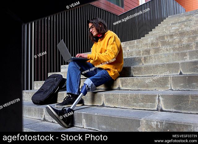 Disabled young man with leg prosthesis using laptop while sitting on steps