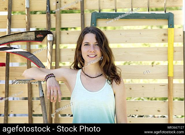 Smiling young woman with gardening equipment standing against fence