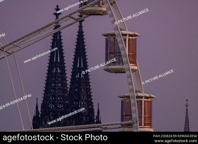 24 August 2023, North Rhine-Westphalia, Cologne: In the foreground are cabins of a Ferris wheel in the morning light, in the background are the two cathedral...