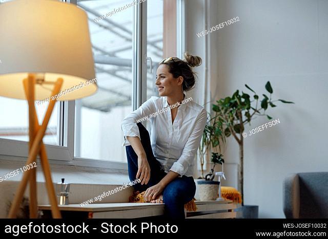 Smiling woman sitting on widow sill looking out from home