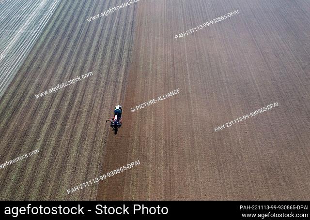 13 November 2023, Mecklenburg-Western Pomerania, Wessin: A tractor pulls a seed drill across a field and sows seed. (Aerial shot with a drone) Farmers in...
