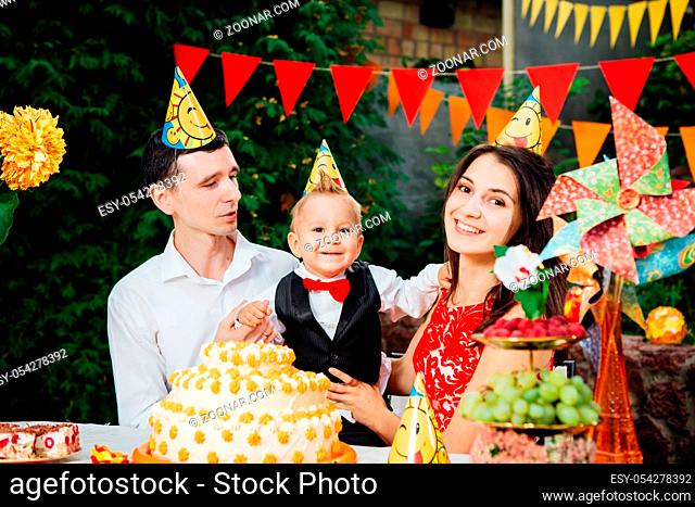 Children birthday theme. family of three Caucasian people sitting in backyard of the house at a festive decorated table in funny hats and caps on their heads