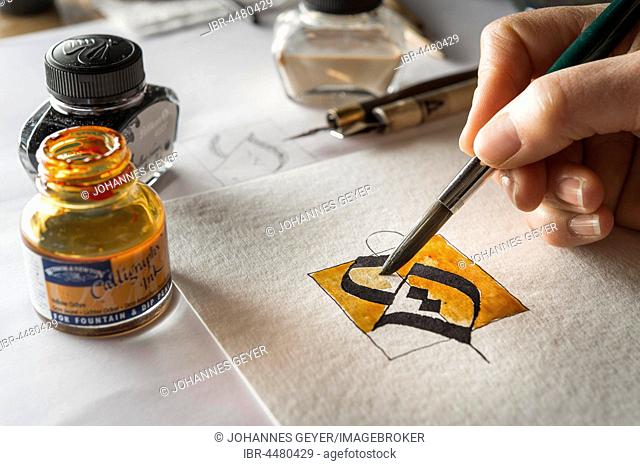 Calligraphy studio, hand paints surface with brush, letter S on torchon paper, inkwells and fountain pen at back, Seebruck, Upper Bavaria, Germany