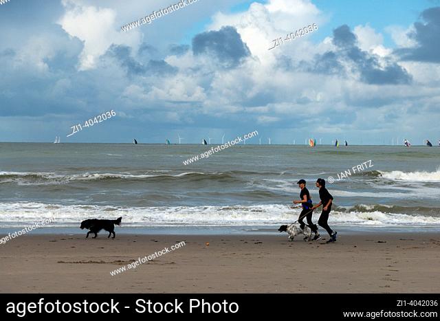 two women running with dogs at the beach and dark clouds above sea with sailboats in Holland