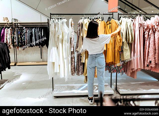 Cute woman choosing elegant dress in clothing store. Female person shopping in fashion boutique, shopaholic, shopper looking on garment