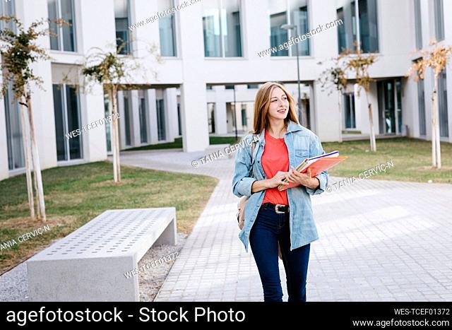 Smiling beautiful blond female student looking away while walking at university campus