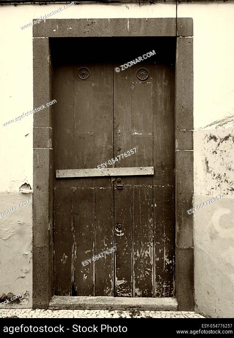 a sepia monochrome image of an old weathered wooden door nailed shut with a wooden bar and flaking paint on an outside wall
