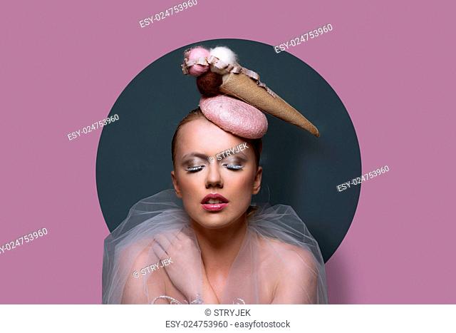 Fine art portrait of a gorgeous woman in fascinator hat in the shape of an ice cream cone with creative makeup and filmy gauze around her shoulders over a...