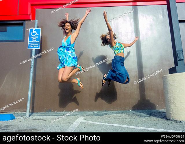 Excited female friends jumping with joy against brown wall at parking lot