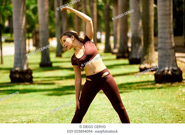 Young blond woman warming up before starting her daily sport routine. The girl does arms stretching in park, near trees. Three quarter length, front view shot