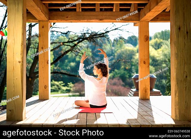 Attractive girl is engaged in yoga on the wooden terrace on the nature background. She sits in the lotus pose on the red yoga mat and stretches her hands over...