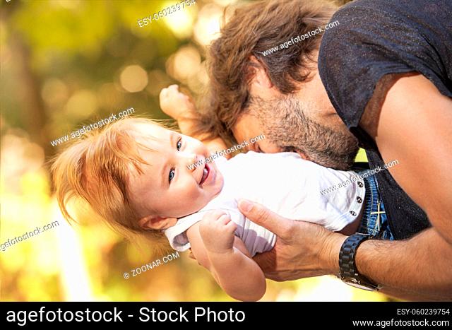 A cute little baby is laughing while his father is biting him playfully outdoors