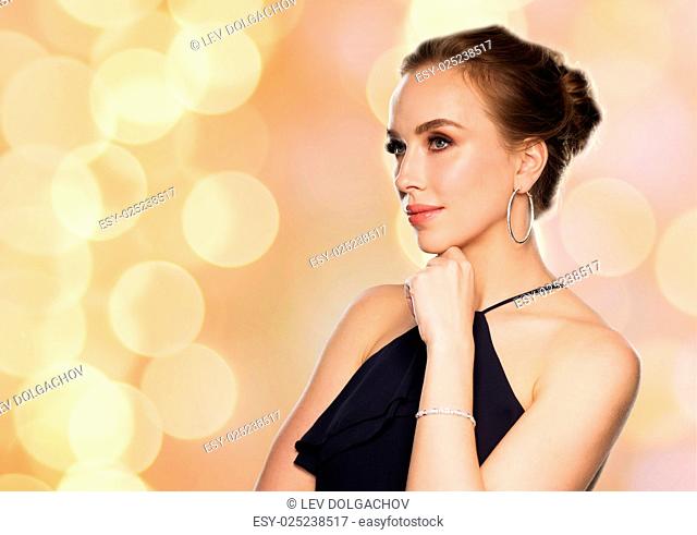 people, luxury, jewelry and fashion concept - beautiful woman in black wearing diamond earrings and bracelet over holidays lights background