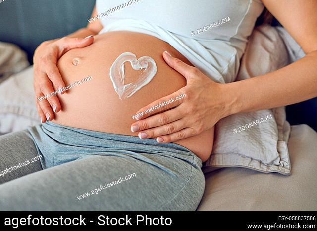 Young pregnant woman drawing a heart in moisturising cream on her baby bump as she cradles her belly in her hands while relaxing on a bed in a close up cropped...