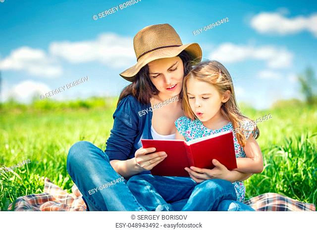 Mother and daughter enjoy reading book sitting on green meadow in the summer park