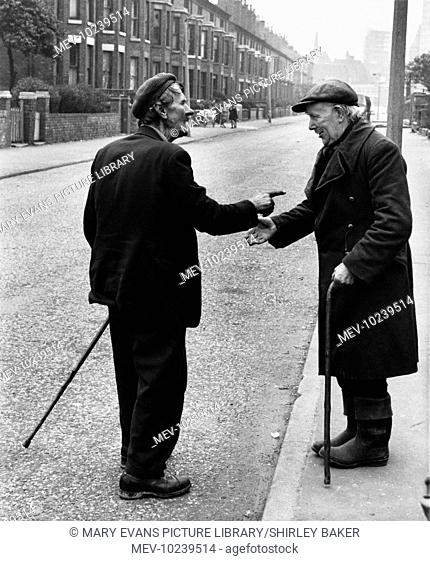 Two slightly scruffy old gentlemen have a seemingly rather frank discussion on a manchester street, Each is carrying a walking stick and wearing flat caps