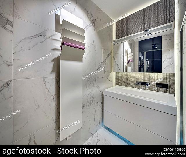 Modern bathroom with light tiles on the walls and the floor. There is a white sink with a chrome faucet and a mirror, a white stand with a towel