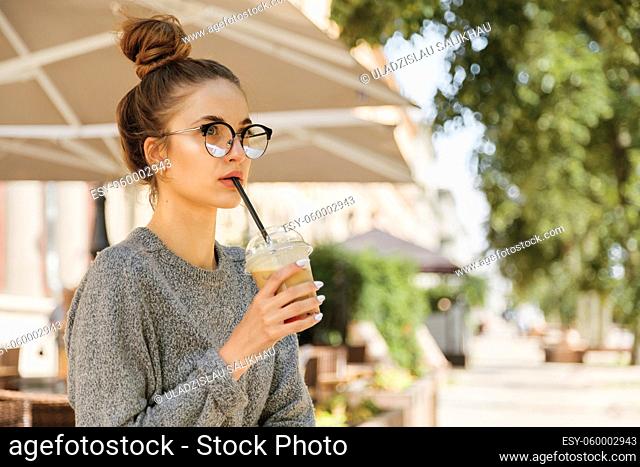 Alone young woman in eyeglasses drinking milkshake and looking away in cozy cafe outdoor
