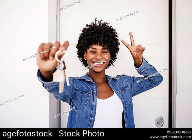 Happy woman showing house keys and peace sign gesture in front of door