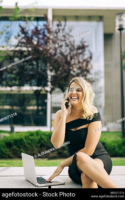 Cheerful businesswoman talking on mobile phone while using laptop outdoors