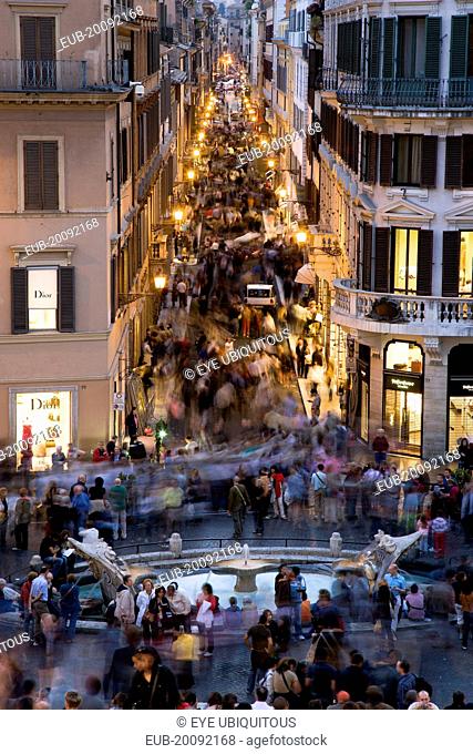 The Via dei Condotti the main shopping street busy with people illuminated at night seen from the Spanish Steps with seated tourists and the Fontana della...