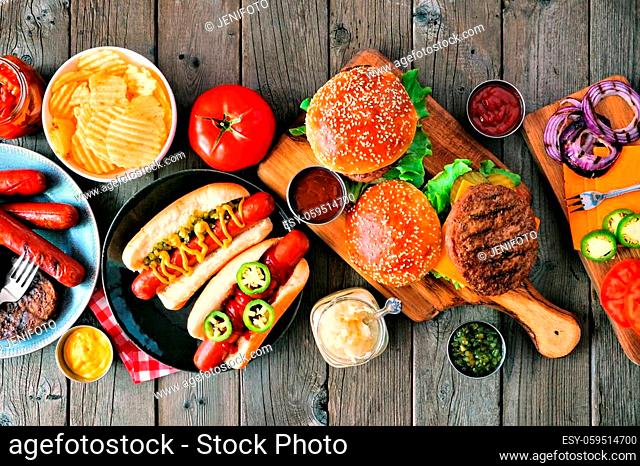 Summer BBQ food table scene with hot dog and hamburger buffet. Above view over a rustic wood background