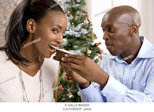 Man feeding woman Christmas cake with Christmas tree in the background