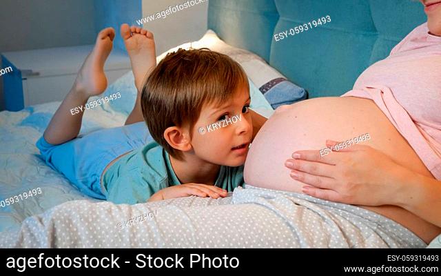 Smiling toddler boy in pajamas talking and hugging unborn baby in big belly of pregnant mother lying in bed at night
