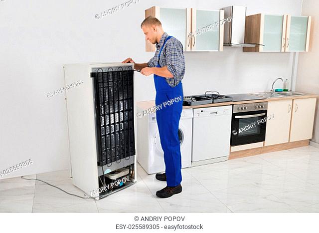 Young Repairman In Overall Repairing Refrigerator In Kitchen Room