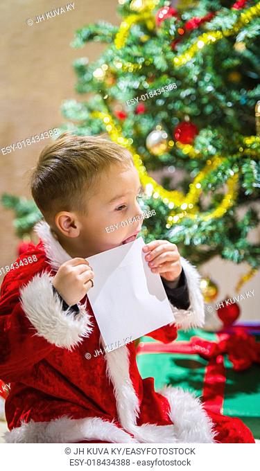Adorable 5 year old boy closing a letter for Santa Claus, Christmas tree and gifts on background