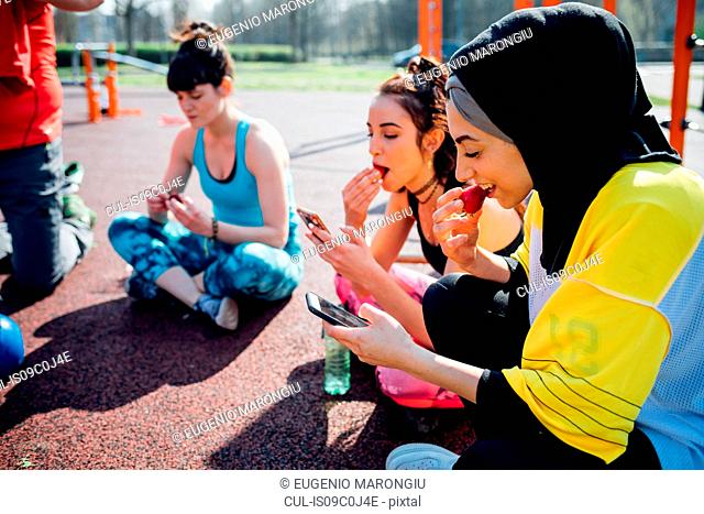 Calisthenics class at outdoor gym, young women sitting looking at smartphones and eating fruit