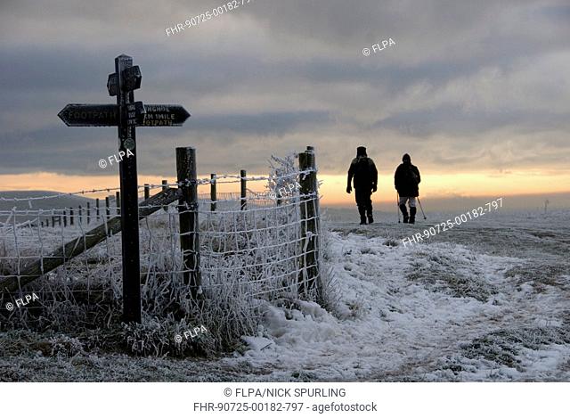 Walkers on frost and snow covered footpath, beside signpost and fence at sunset, Ridgeway Path, near Pitstone Hill, Chilterns, Buckinghamshire, England