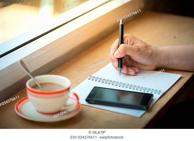 Male right hand writing on notebook with smart phone and coffee cup on wood bar beside window in cafe in morning time