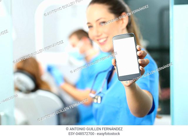 Stomatologist showing phone screen and looking at you in a dentist office interior with a doctor working in the background