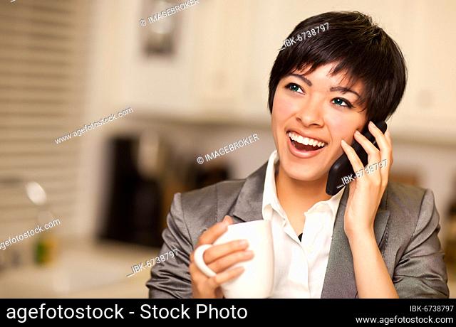 Pretty smiling multiethnic woman with coffee and talking on a cell phone in her kitchen