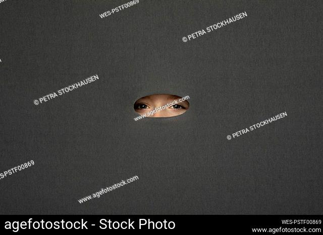 Eyes of teenage girl peeking through hole in gray background