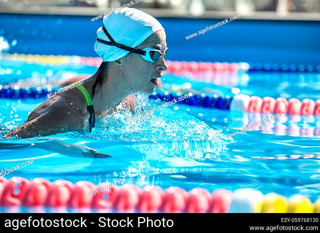 Emotional close-up photo of female swimmer in the motion in the swimming pool outdoors. She wears a black-lime swimsuit, a white swim cap and swim glasses