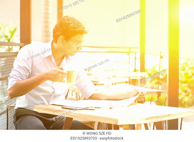 Asian Indian business man reading newspaper while drinking a cup hot milk tea at cafeteria, with beautiful golden sunlight