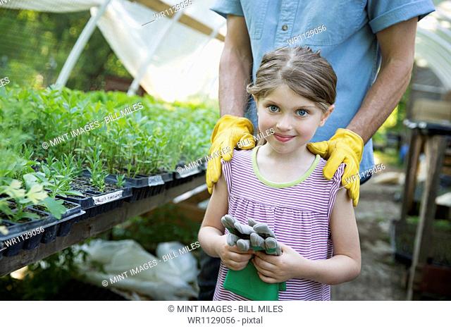 Outdoors in summer. On the farm. Children and adults working together. A man and a young child with gardening gloves, standing beside a bench of young seedling...