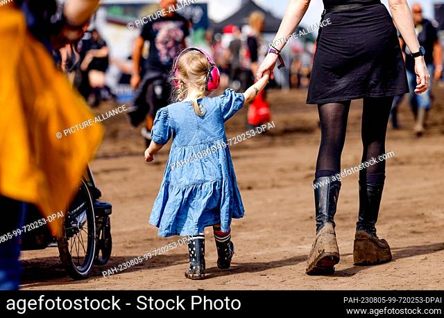 05 August 2023, Schleswig-Holstein, Wacken: A little girl wearing hearing protection walks around the festival grounds at the Wacken Open Air holding her...