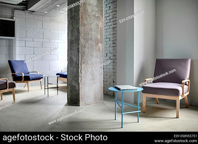 Work zone in the office in a loft style with a concrete column and light walls. Near the column there are multi-colored armchairs with wooden legs and small...