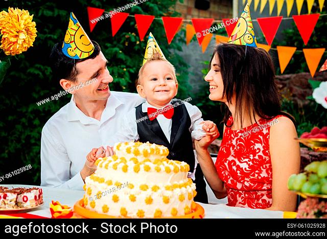 Children birthday theme. family of three Caucasian people sitting in backyard of the house at a festive decorated table in funny hats and caps on their heads